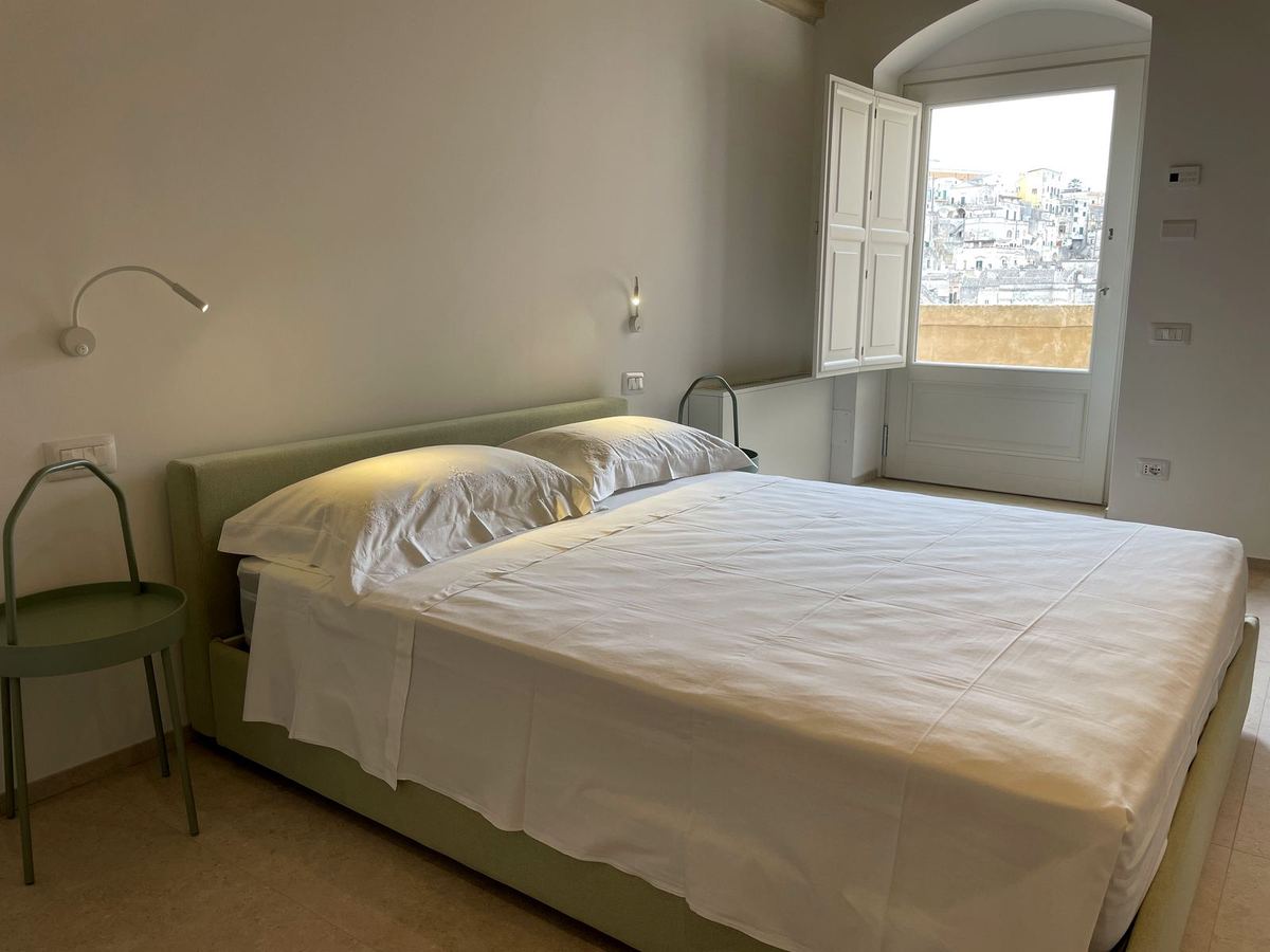 Standard Suite: Doppelbett mit salbeigrünem Kopfteil und geöffnetem Fenster zu den Sassi